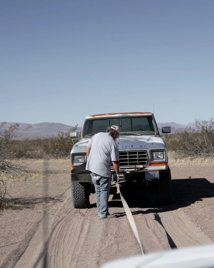A driver rigging a recovery strap to a pickup on a dirt road